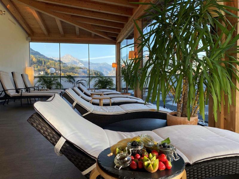 Terrasse mit bequemen Liegestühlen, großen Pflanzen und Panoramablick auf Berge und See.