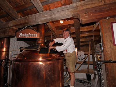 Muž v tradičním kroji stojí vedle velkého měděného kotle v rustikálním pivovaru.