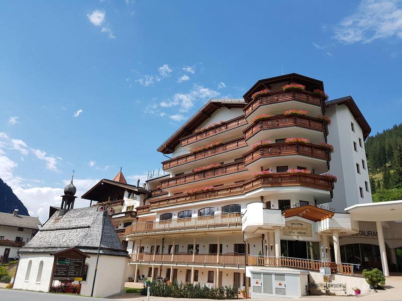 Mehrstöckiges alpines Hotel mit Balkonen, weißem Anbau und dahinter Bergen unter blauem Himmel.