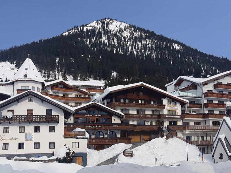 Winterszene eines verschneiten Bergdorfes mit Holzhäusern und Tannen am Hang unter blauem Himmel.