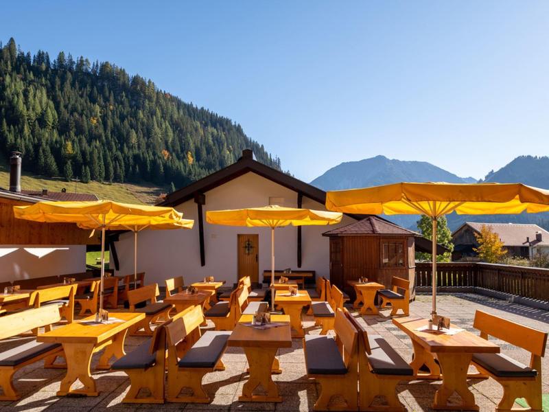 Sonnige Terrasse mit Holztischen und gelben Sonnenschirmen vor Bergen und blauem Himmel.