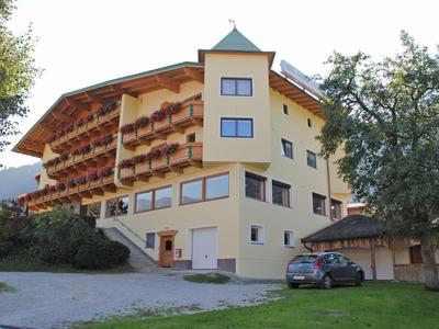 Großes gelbes Gebäude mit Balkonen, Blumen, Bäumen und einem geparkten Auto davor.