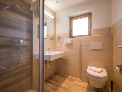 Kleines Badezimmer mit Dusche, Waschbecken, Toilette und Fenster, beige Fliesen und Holzoptik-Boden.