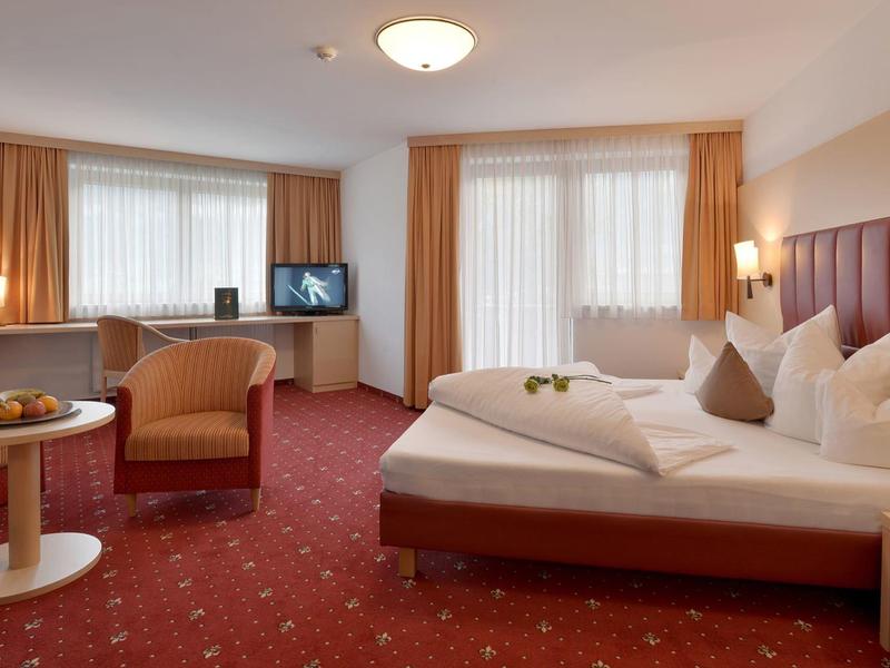 Gemütliches Hotelzimmer mit rotem Teppich, Doppelbett, zwei Sesseln, Tisch und großen Fenstern.