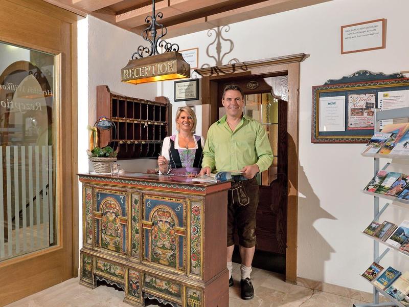 Zwei Personen in traditioneller Kleidung stehen an einem verzierten Holzschalter in einem hellen Raum.