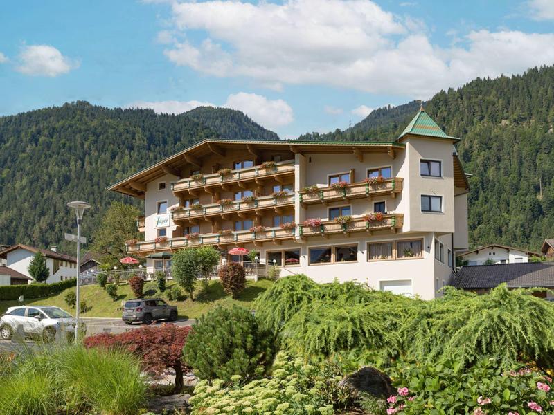Großes Wohnhaus mit Balkonen, grünen Hecken und Bergen im Hintergrund unter blauem Himmel.