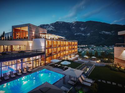 Moderne Hotelanlage mit beleuchtetem Pool, Liegestühlen und Bergblick bei Sonnenuntergang.