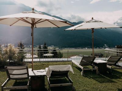 Sonnenliegen mit weißen Sonnenschirmen auf einer Rasenfläche mit Bergblick und See im Hintergrund.