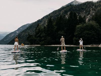 Drei Personen auf Stand-Up-Paddle-Boards auf einem See vor bewaldeten Bergen.