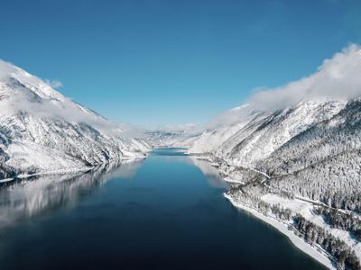 Blick auf einen ruhigen, schmalen See umgeben von schneebedeckten Bergen unter klarem Himmel.