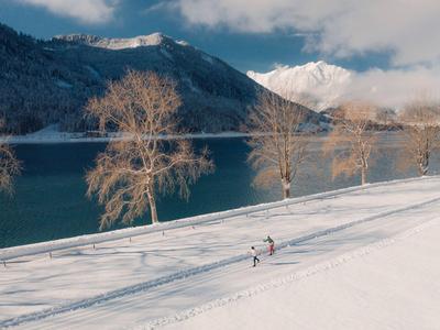 Schneebedeckte Landschaft mit See, Bergen und Menschen beim Langlaufen.