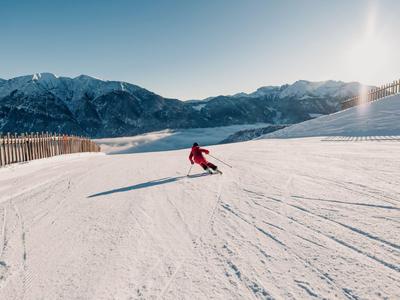 Skifahrer fährt auf breiter, schneebedeckter Piste vor sonnigen Berggipfeln.