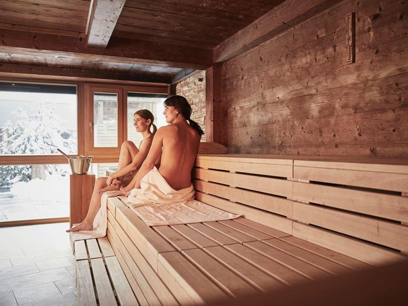 Zwei Personen sitzen auf Holzbänken in einer warm beleuchteten Sauna mit Fensterfront.