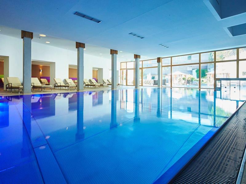 Modernes Hallenbad mit blauem Wasser, Liegen und großen Fenstern für Tageslicht.