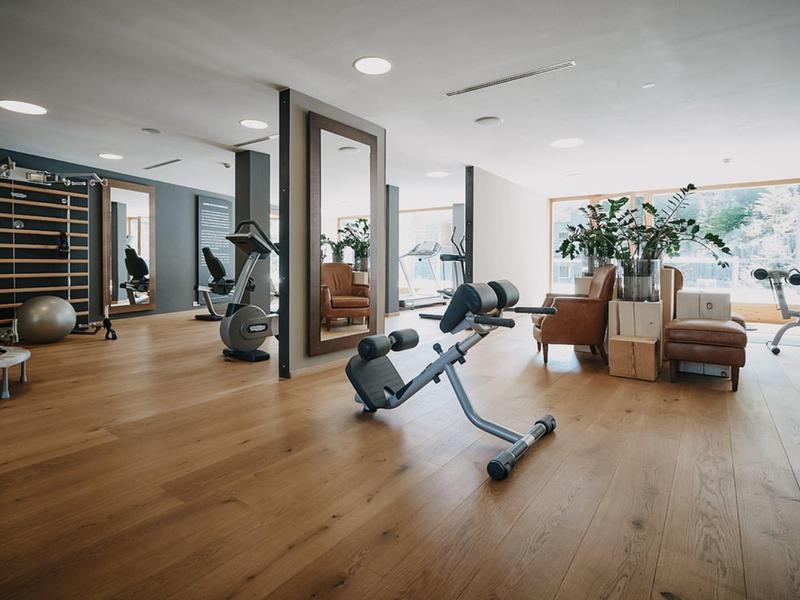 Heller Fitnessraum mit Holzfußboden, Trainingsgeräten, Spiegeln und großen Fenstern.