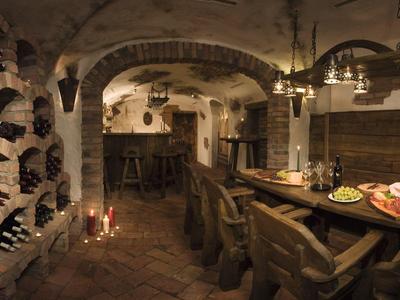 Rustikaler Weinkeller mit Steinwänden, Holzregalen und einem Tisch mit Speisen und Holzstühlen.