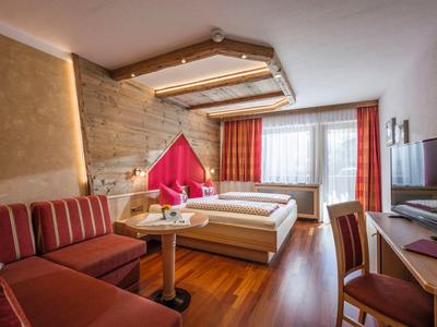 Gemütliches Hotelzimmer mit zwei Betten, Sofa, Holzboden und großer Fensterfront mit roten Vorhängen.