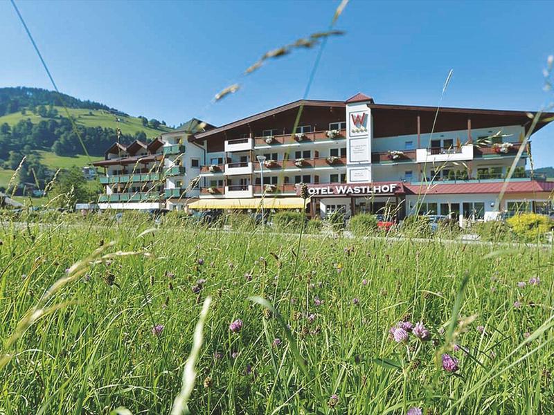Hotelgebäude mit Balkonen vor einer grünen Wiese und Hügeln im Hintergrund