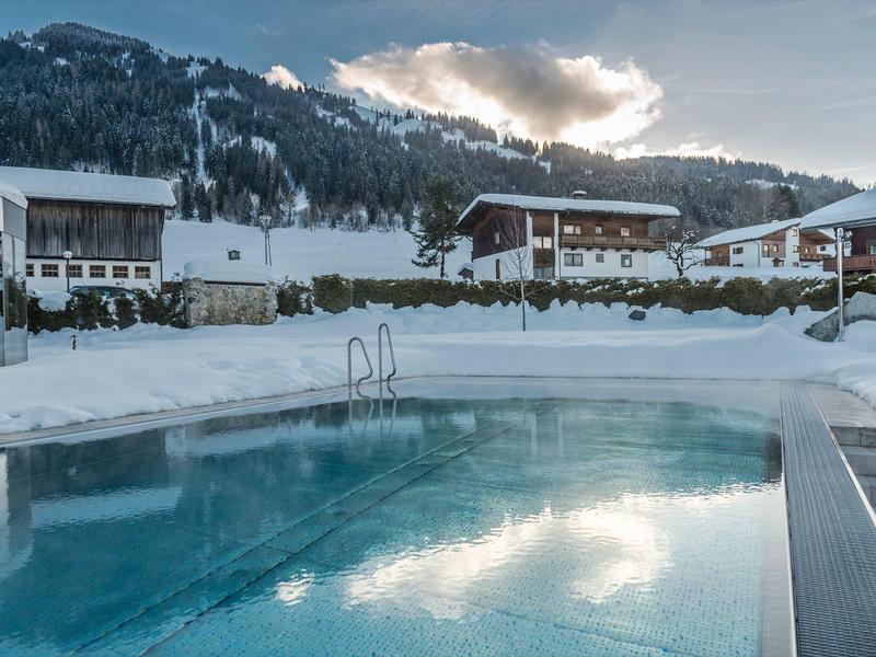 Beheizter Außenpool mit Schnee und Bergen im Hintergrund, umgeben von Chalet-Häusern.