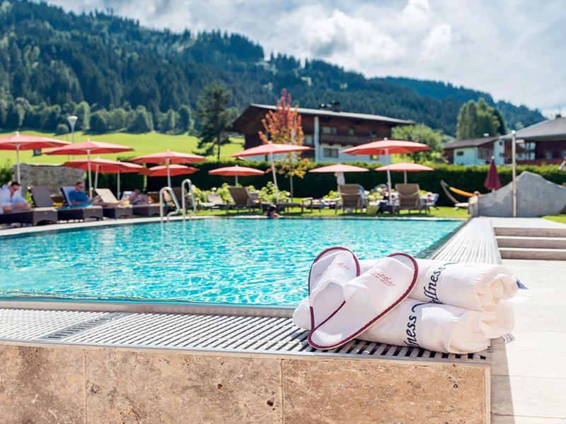 Schwimmbad mit Herz-Sonnenbrillen am Beckenrand und Sonnenschirmen vor bergiger Kulisse.