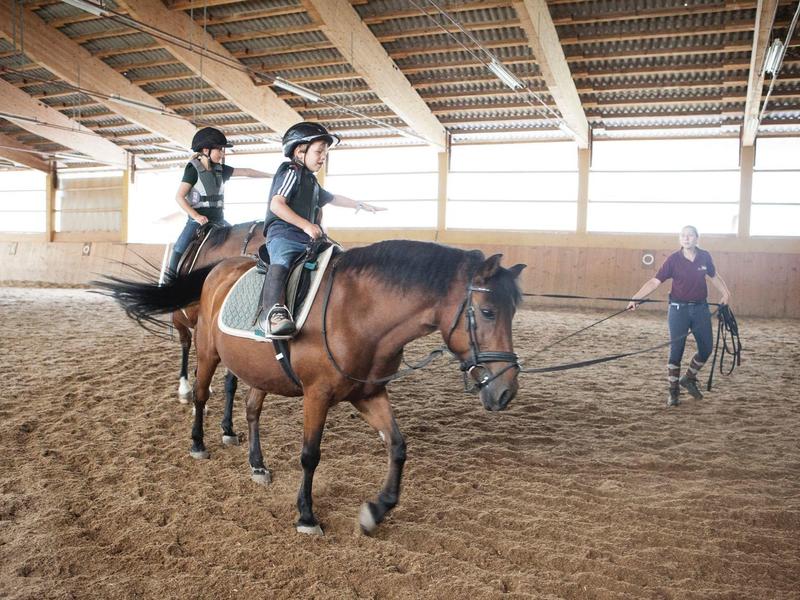 Zwei Kinder reiten in einer Reithalle, begleitet von einer erwachsenen Person beim Longieren.