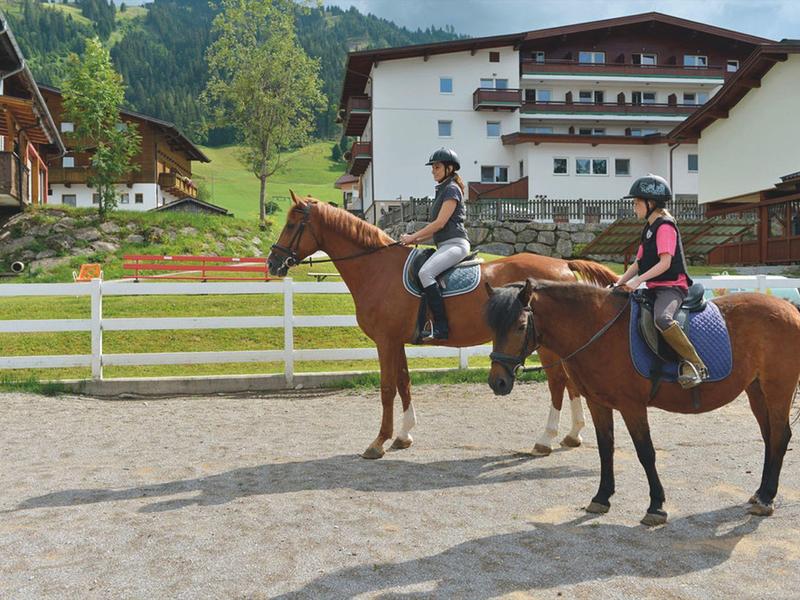 Zwei Personen reiten auf Pferden in einem umzäunten Bereich vor Alpenhäusern.