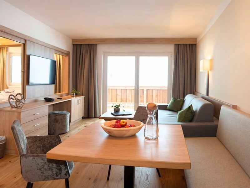 Helles Wohnzimmer mit Sofa, Esstisch, Fernseher und großen Fenstern mit Vorhängen.