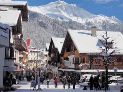 Winterliches Dorf mit verschneiten Häusern und Bergen, blauem Himmel und Spaziergängern auf der Straße.