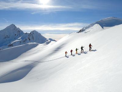 Fünf Bergsteiger wandern im Schnee über einen sonnigen, schneebedeckten Berghang mit blauem Himmel.