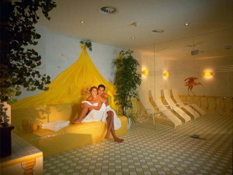 Romantisches Spa-Zimmer mit zwei Personen auf gelber Liege, grünen Pflanzen und weißen Liegestühlen.