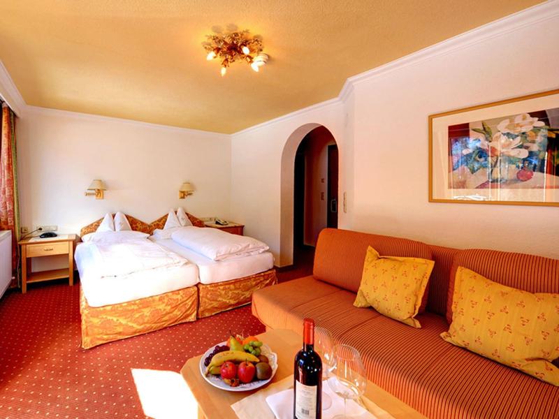 Gemütliches Hotelzimmer mit zwei Einzelbetten, oranger Couch, Obstkorb und Weinflasche, warmes Licht.