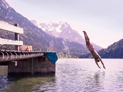 Person springt vom Holzsteg in ruhigen Bergsee mit Bergen und wolkenverhangenem Himmel im Hintergrund.