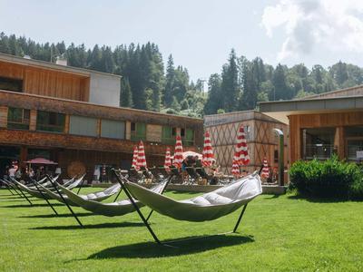 Grüne Wiese mit Hängematten vor einem zweistöckigen Holzgebäude, umgeben von Nadelbäumen.