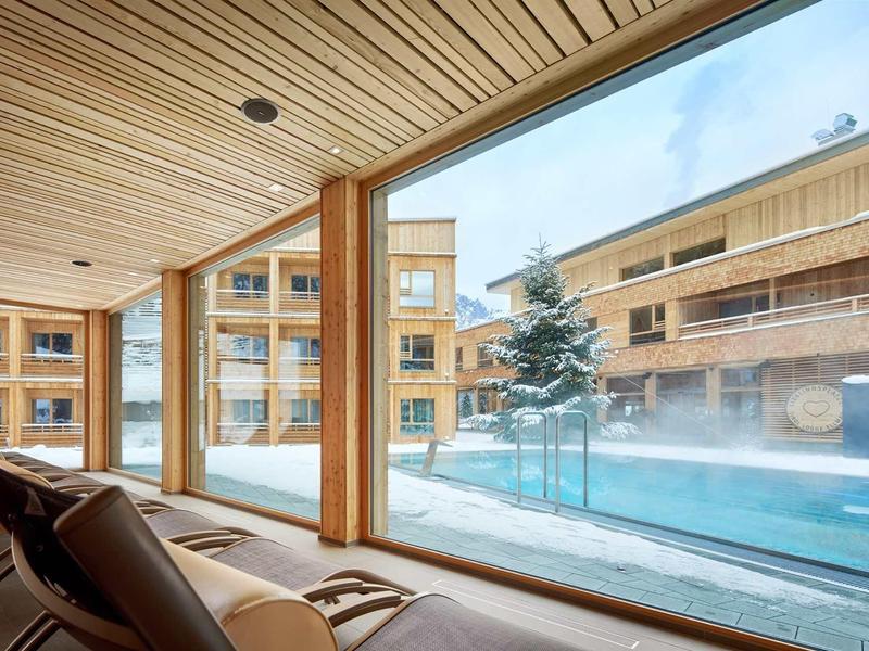 Blick aus einem gemütlichen Holzraum auf verschneite Innenhof mit Tannen und modernes Gebäude.