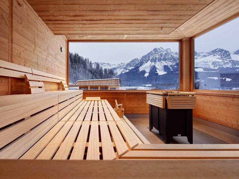Moderne Sauna mit Holzinterieur und großer Panoramafensteransicht auf verschneite Berge.