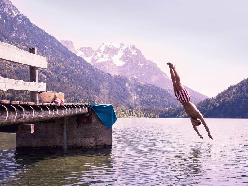 Person springt vom Holzsteg in ruhigen Bergsee mit Bergen und wolkenverhangenem Himmel im Hintergrund.