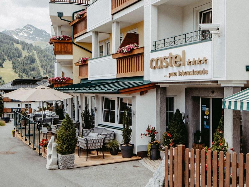 Außenansicht eines weißen Hotels mit Holzbalustern, Blumentöpfen und Bergen im Hintergrund.