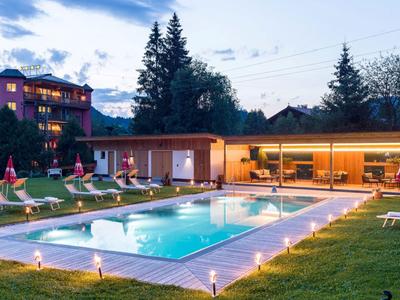 Illuminated pool area with lounge chairs and modern buildings in the background at dusk.