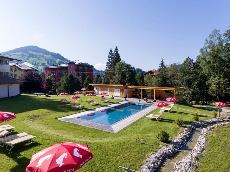 Schwimmbad mit grüner Liegewiese, roten Sonnenschirmen und umliegenden Bäumen unter klarem Himmel.