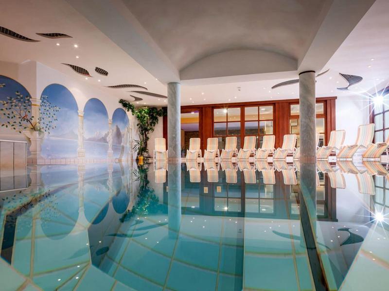 Innenbereich eines Schwimmbads mit klarem Wasser, Säulen und weißen Liegestühlen an der Seite.