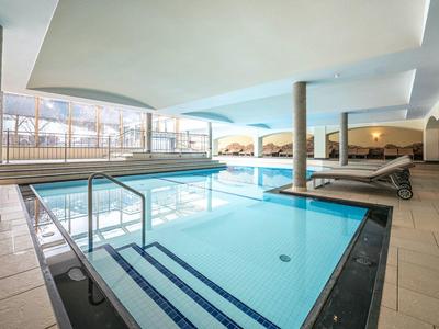 Modernes Hallenbad mit großem Schwimmbecken, hellem Boden, Säulen und mehreren Liegestühlen.