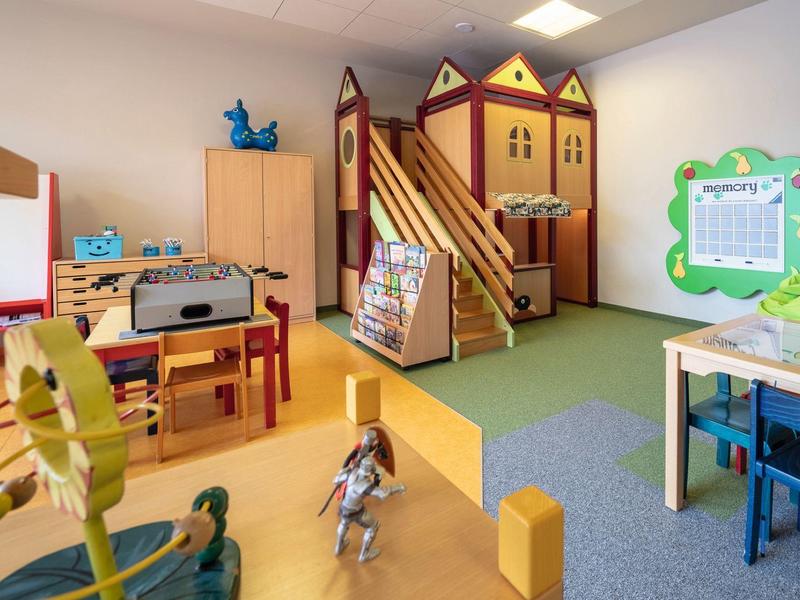 Kindergartenraum mit Spielhaus, Spielzeug, bunten Möbeln und Lernmaterialien in hellem Licht.