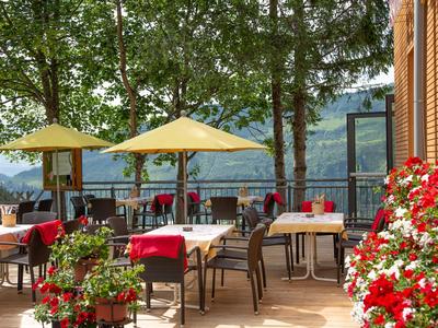 Sonnige Terrasse mit Tischen, Stühlen und gelben Sonnenschirmen, umgeben von Blumen und Bäumen.