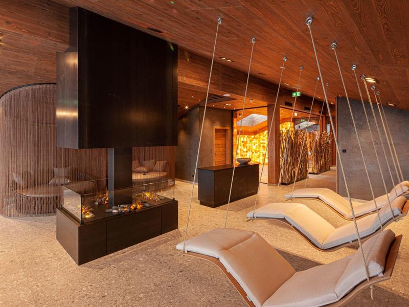 Moderne Lounge mit Holzdecke, zwei hängenden Liegen und zentralem Kamin in warmem Licht.