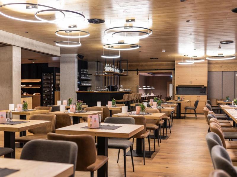 Moderner Speisesaal mit Holztischen, gepolsterten Stühlen und runden Deckenlampen.