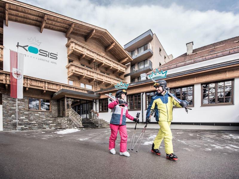 Zwei Kinder in bunten Skibekleidungen laufen mit Skiern vor alpinen Holzhäusern bei bewölktem Himmel.