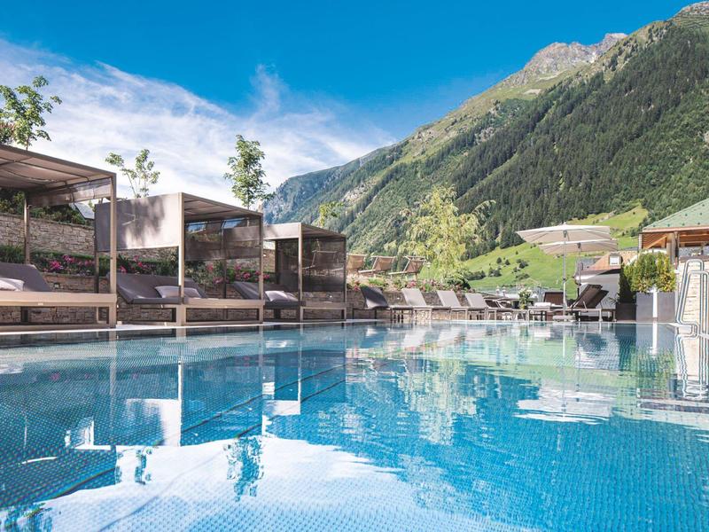 A hotel pool with lounge chairs and mountain scenery in the background under clear sky.
