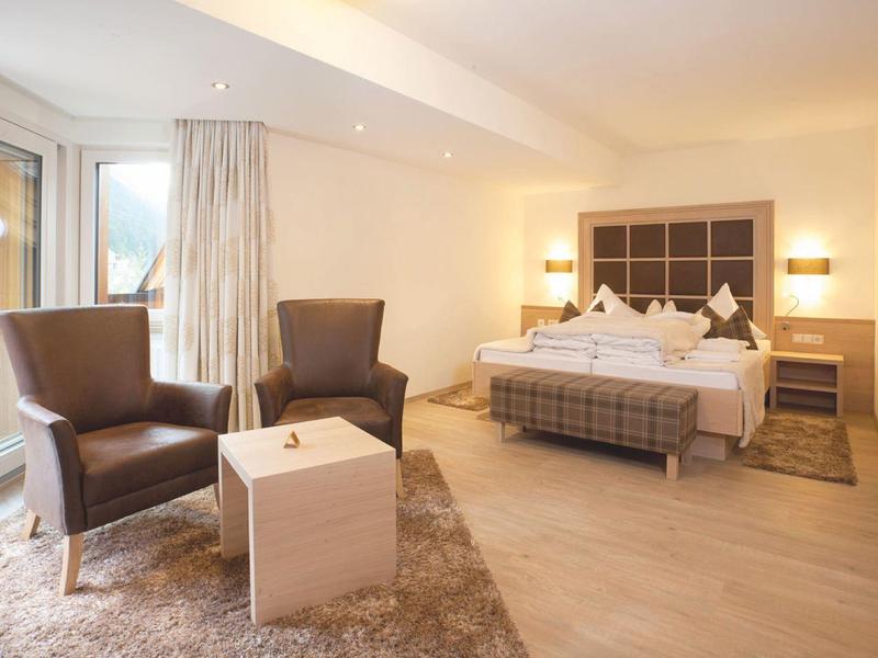 Bright hotel room with two armchairs, bed, and small table on a rug.