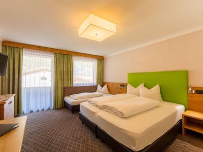 Helles Hotelzimmer mit grünem Kopfteil, Doppel- und Einzelbett, TV und großem Fenster mit grünen Vorhängen.