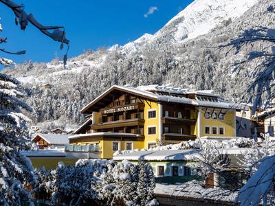 Geel hotel voor met sneeuw bedekte bergen en bomen onder een blauwe lucht.